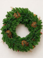 Load image into Gallery viewer, A green artificial pine wreath with natural brown pinecones, small red berries, and a thin metal circular frame, shown against a plain off-white background.